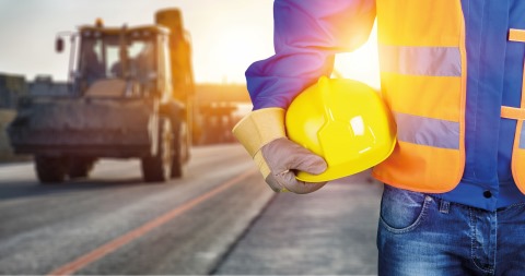 Links steht ein Bagger im Hintergrund, rechts im Vordergrund der Torso eines Mannes in Baustellenkleidung, mit einem gelben Helm in der Hand.