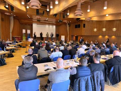 Im Vordergrund sind Stuhlreihen besetzt mit nach vorne blickenden Menschen zu sehen. An der Saalr&uuml;ckwand im Bildhintergrund stehen 5 Personen auf der B&uuml;hne und diskutieren mit dem Publikum.
