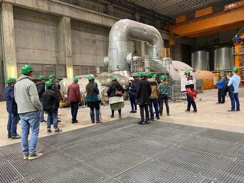 In einem gro&szlig;en Kraftwerksraum steht eine Gruppe von Menschen mit gr&uuml;nen Schutzhelmen vor einer Pumpenanlage.