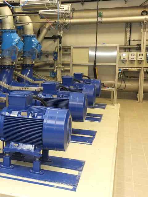 Raum in einem Betriebsgeb&auml;ude einer abwassertechnischen Anlage. Es sind mehrere gro&szlig;e blaue Pumpen zu sehen, die links auf dem Boden befestigt sind. An den W&auml;nden verlaufen silberfarbene Rohre.