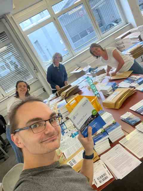Ein Raum mit Fenstern am hinteren Ende. Im Vordergrund Florian Wesche vor einem gro&szlig;en Tisch, auf dem die einzelnen Bestandteile f&uuml;r den Versadn sowie viele B&uuml;cher in Stapeln sortiert liegen. Im Hintergrund schauen v.l.n.r. Sabine Schaible-Friedel, Christiane Sch&auml;fer und Simone Schneider in die Kamera.