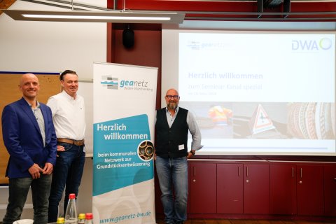 Links in einem Vortragsraum stehen drei Referenten um ein Roll-Up von geanetz.plus gruppiert. Rechts ist eine Leinwand mit einer Begr&uuml;&szlig;ungsfolie zum Seminar zu sehen.