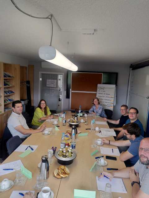Die Teilnehmenden des Seminars sitzen um einen ovalen Konferenztisch und schauen in die Kamera. Die Seminarleiterin sitzt am Ende des Tischs vor einem Flipchart.