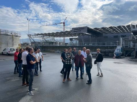 Einige Teilnehmende des Treffens der Bodensee-Nachbarschaft stehen w&auml;hrend der Exkursion auf einem offenen Platz der ARA Morgental. Im Hintergrund das Solarfaltdach und weitere Anlagenteile unter teils bew&ouml;lktem, blauem Himmel. 