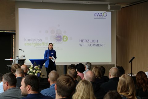 Die Moderatorin Hannah Pinell er&ouml;ffnet auf der B&uuml;hne den Kongress. Im Vordergrund ist der vollbesetzte Zuschauerraum zu sehen, im Hintergrund die Er&ouml;ffnungsfolie auf einer Leinwand.