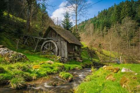 Berglandschaft mit einer H&uuml;tte an einem Bach, umgeben von gr&uuml;nen Wiesen und W&auml;ldern im Hintergrund.